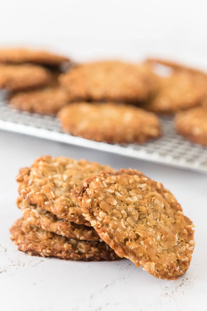 pile of anzac biscuit cookies in front of cooling rack piled with more cookies