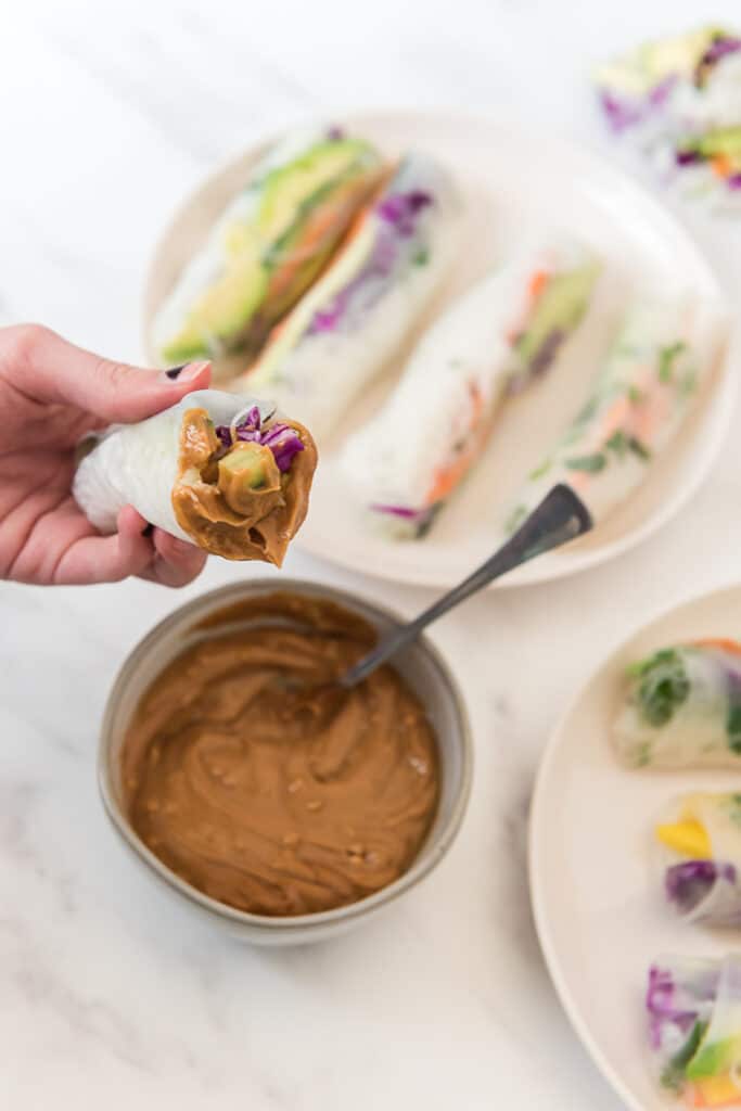 child's hand holding cut spring roll that has been dipped in peanut sauce over platter of more spring rolls and bowl of sauce