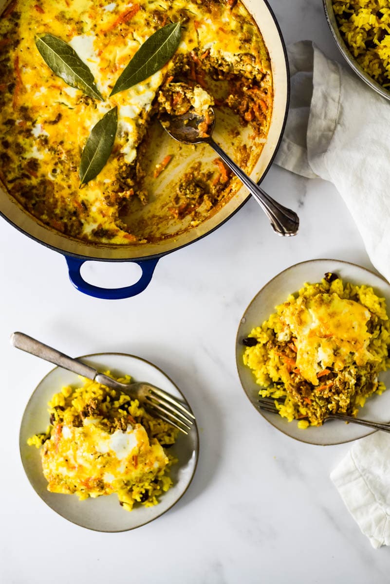 two plates piled with bobotie with forks on the side, next to serving dish with spoon