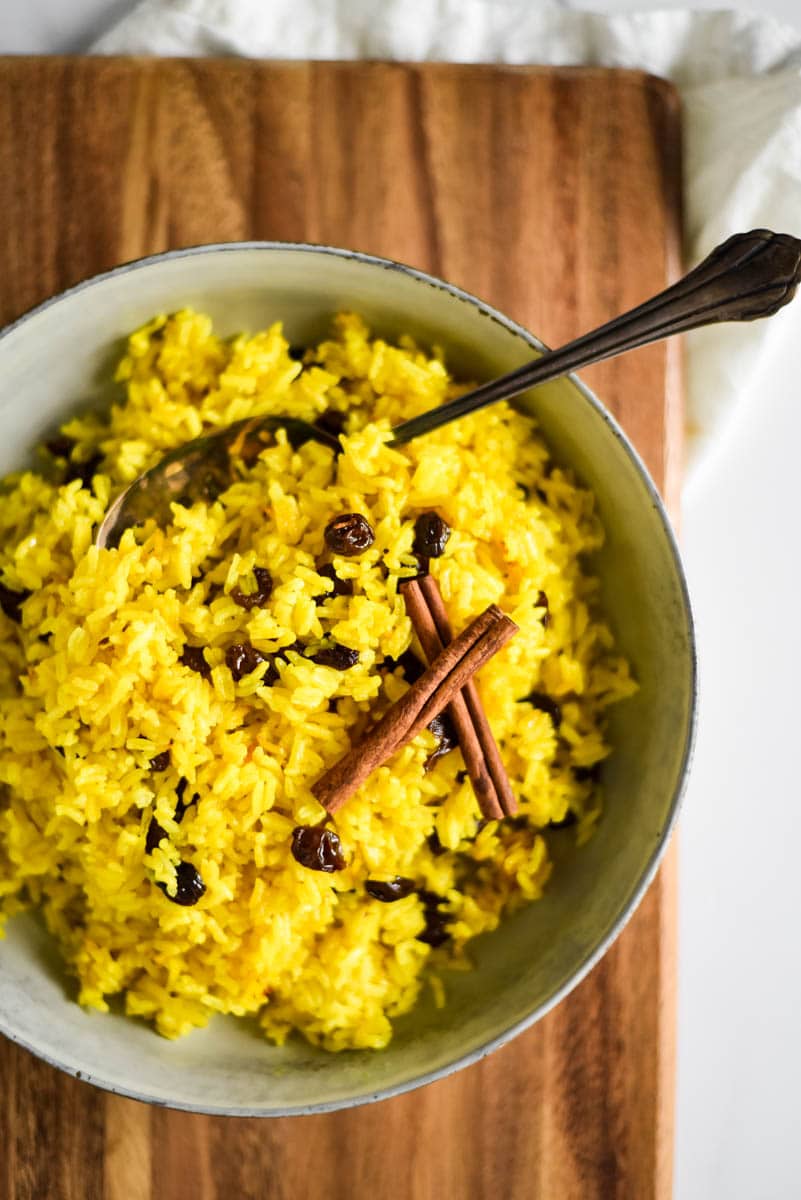 ceramic bowl filled with south african yellow rice. Serving spoon and two crossed cinnamon sticks are on the top of the rice. Bowl is sitting on wooden cutting board
