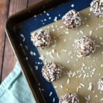 Chocolate coconut ball cookies on a baking sheet with a silicone baking mat. There is coconut sprinkled all around the baking mat. It is all on a wooden table